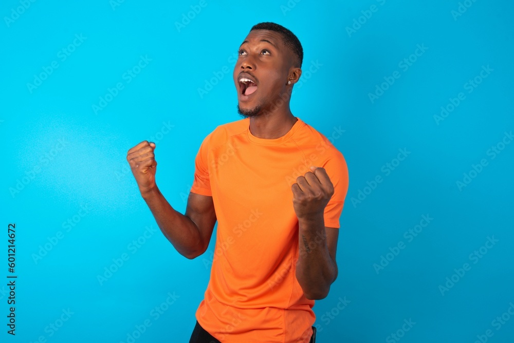 Handsome man wearing orange T-shirt over blue background celebrating surprised and amazed for success with arms raised and eyes closed. Winner concept. Handsome man wearing orange T-shirt over blue background celebrating surprised and amazed for success with arms raised and eyes closed. Winner concept.