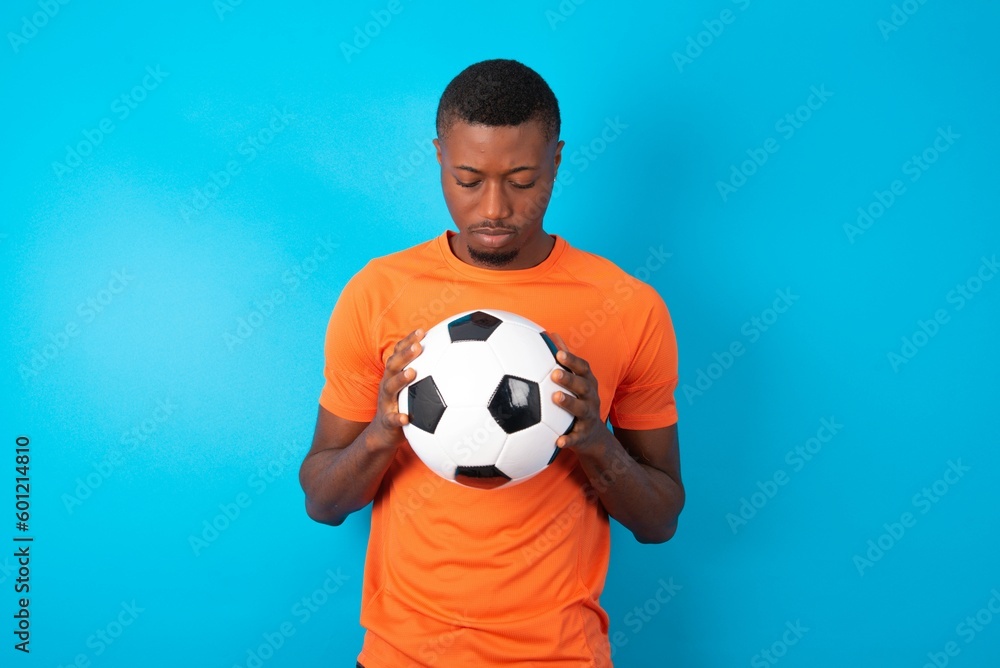 Sad Man wearing orange T-shirt holding a ball over blue background desperate and depressed with tears on her eyes suffering pain and depression in sadness facial expression and emotion concept Sad Man wearing orange T-shirt holding a ball over blue background desperate and depressed with tears on her eyes suffering pain and depression in sadness facial expression and emotion concept