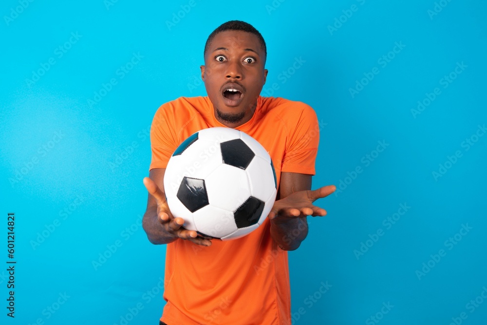 Frustrated Man wearing orange T-shirt holding a ball over blue background feels puzzled and hesitant, shrugs shoulders in bewilderment, keeps mouth widely opened, doesn't know what to do. Frustrated Man wearing orange T-shirt holding a ball over blue background feels puzzled and hesitant, shrugs shoulders in bewilderment, keeps mouth widely opened, doesn't know what to do.