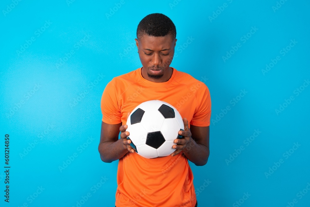 Man wearing orange T-shirt holding a ball over blue background Tired hands covering face, depression and sadness, upset and irritated for problem Man wearing orange T-shirt holding a ball over blue background Tired hands covering face, depression and sadness, upset and irritated for problem