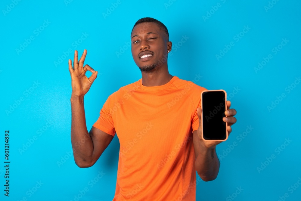 Handsome man wearing orange T-shirt over blue background holding in hands cell showing ok-sign Handsome man wearing orange T-shirt over blue background holding in hands cell showing ok-sign