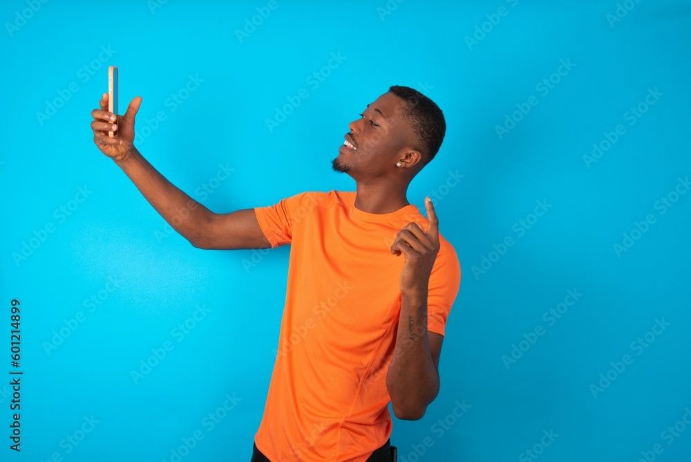 Handsome man wearing orange T-shirt over blue background smiling and taking a selfie ready to post it on her social media. Handsome man wearing orange T-shirt over blue background smiling and taking a selfie ready to post it on her social media.