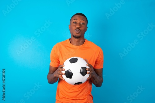 Shot of pleasant looking Man wearing orange T-shirt holding a ball over blue background , pouts lips, looks at camera, Human facial expressions © Jihan