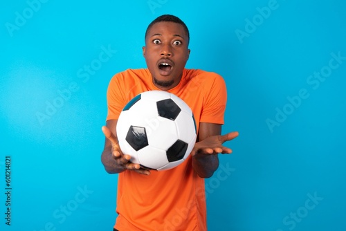 Frustrated Man wearing orange T-shirt holding a ball over blue background feels puzzled and hesitant, shrugs shoulders in bewilderment, keeps mouth widely opened, doesn't know what to do. © Jihan