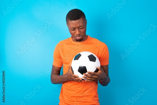 Dismal gloomy rejected Man wearing orange T-shirt holding a ball over blue background has problems and difficulties, curves lower lip and closes eyes in despair, being in depression