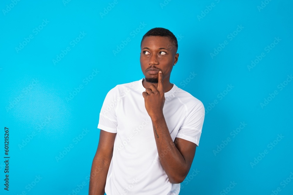 Lovely dreamy Handsome man wearing white T-shirt over blue background keeps finger near lips looks aside copy space. Lovely dreamy Handsome man wearing white T-shirt over blue background keeps finger near lips looks aside copy space.