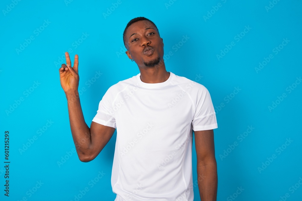 Handsome man wearing white T-shirt over blue background makes peace gesture keeps lips folded shows v sign. Body language concept Handsome man wearing white T-shirt over blue background makes peace gesture keeps lips folded shows v sign. Body language concept
