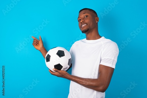 Optimistic Man wearing white T-shirt holding a ball over blue background points with both hands and  looking at empty space.