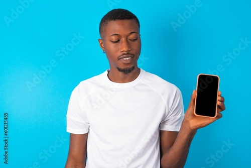 Photo of astonished crazy Handsome man wearing white T-shirt over blue background hold smartphone dislike feedback concept