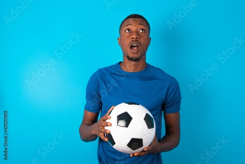 Young man wearing sport T-shirt holding a ball over blue background yawns with opened mouth stands. Daily morning routine