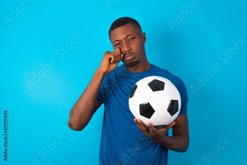 Disappointed dejected Young man wearing sport T-shirt holding a ball over blue background wipes tears stands stressed with gloomy expression. Negative emotion