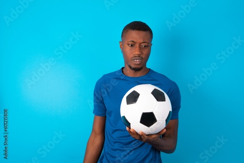 Portrait of dissatisfied Young man wearing sport T-shirt holding a ball over blue background smirks face, purses lips and looks with annoyance at camera, discontent hearing something unpleasant