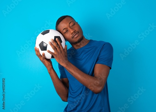 Young man wearing sport T-shirt holding a ball over blue background leans on pressed palms closes eyes and has pleasant smile dreams about something