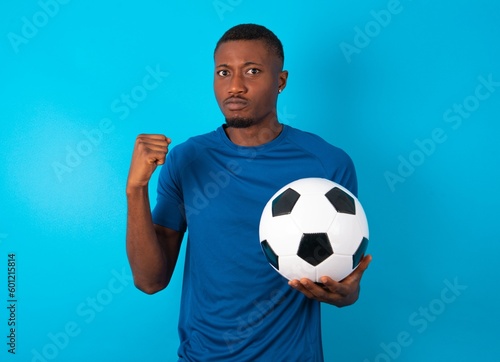 Irritated Young man wearing sport T-shirt holding a ball over blue background blows cheeks with anger and raises clenched fists expresses rage and aggressive emotions. Furious model