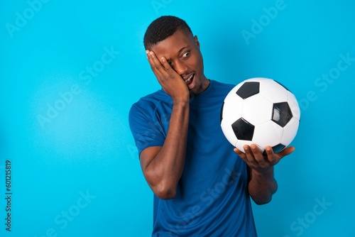 Young man wearing sport T-shirt holding a ball over blue background keeps hands on cheeks has bored displeased expression. Stressed hopeless model