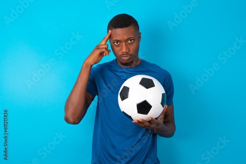 Serious concentrated Young man wearing sport T-shirt holding a ball over blue wall keeps fingers on temples, tries to ease tension, gather with thoughts and remember important information for exam