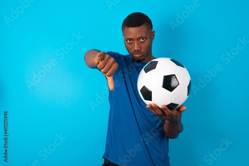 Young man wearing sport T-shirt holding a ball over blue background looking unhappy and angry showing rejection and negative with thumbs down gesture. Bad expression.