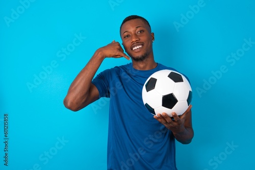 Young man wearing sport T-shirt holding a ball over blue background makes phone gesture, says call me back again, has glad expression.