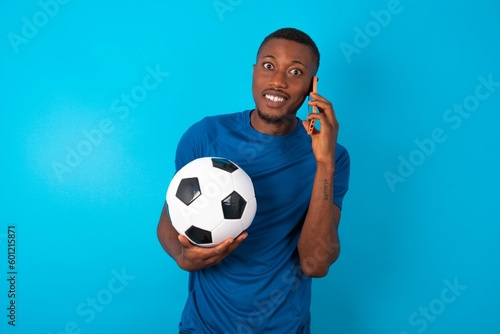 Smiling Young man wearing sport T-shirt holding a ball over blue background talks via cellphone, enjoys pleasant great conversation. People, technology, communication concept