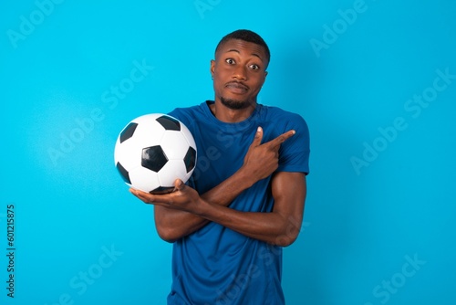 Young man wearing sport T-shirt holding a ball over blue background crosses arms and points at different sides hesitates between two items or variants. Needs help with decision