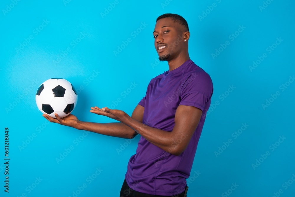 Man wearing purple T-shirt holding a ball over blue background Inviting to enter smiling natural with open hands. Welcome sign. Man wearing purple T-shirt holding a ball over blue background Inviting to enter smiling natural with open hands. Welcome sign.
