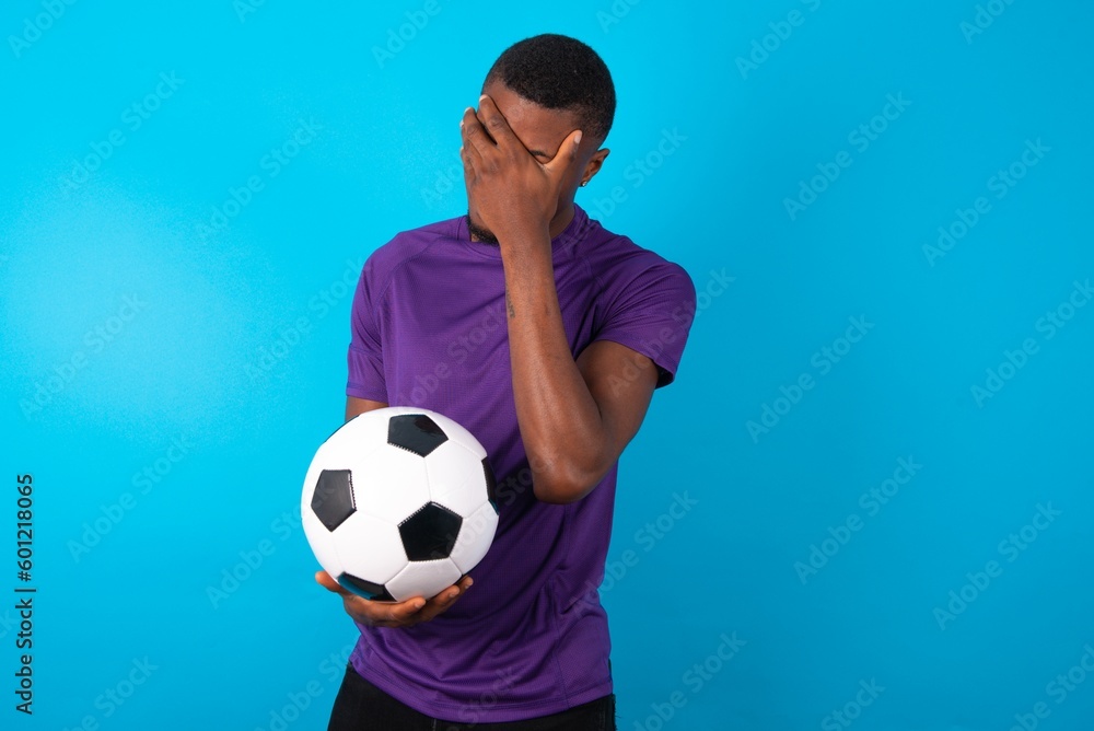 Man wearing purple T-shirt holding a ball over blue background making facepalm gesture while smiling amazed with stupid situation. Man wearing purple T-shirt holding a ball over blue background making facepalm gesture while smiling amazed with stupid situation.