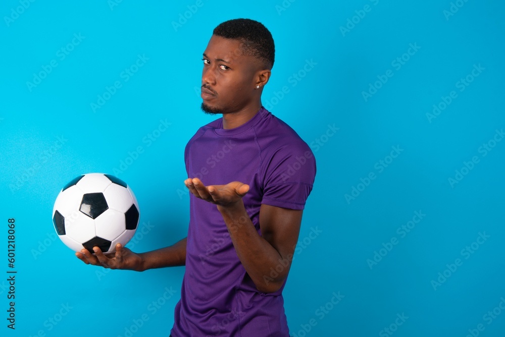 Studio shot of frustrated Man wearing purple T-shirt holding a ball over blue background gesturing with raised palm, frowning, being displeased and confused with dumb question. Studio shot of frustrated Man wearing purple T-shirt holding a ball over blue background gesturing with raised palm, frowning, being displeased and confused with dumb question.