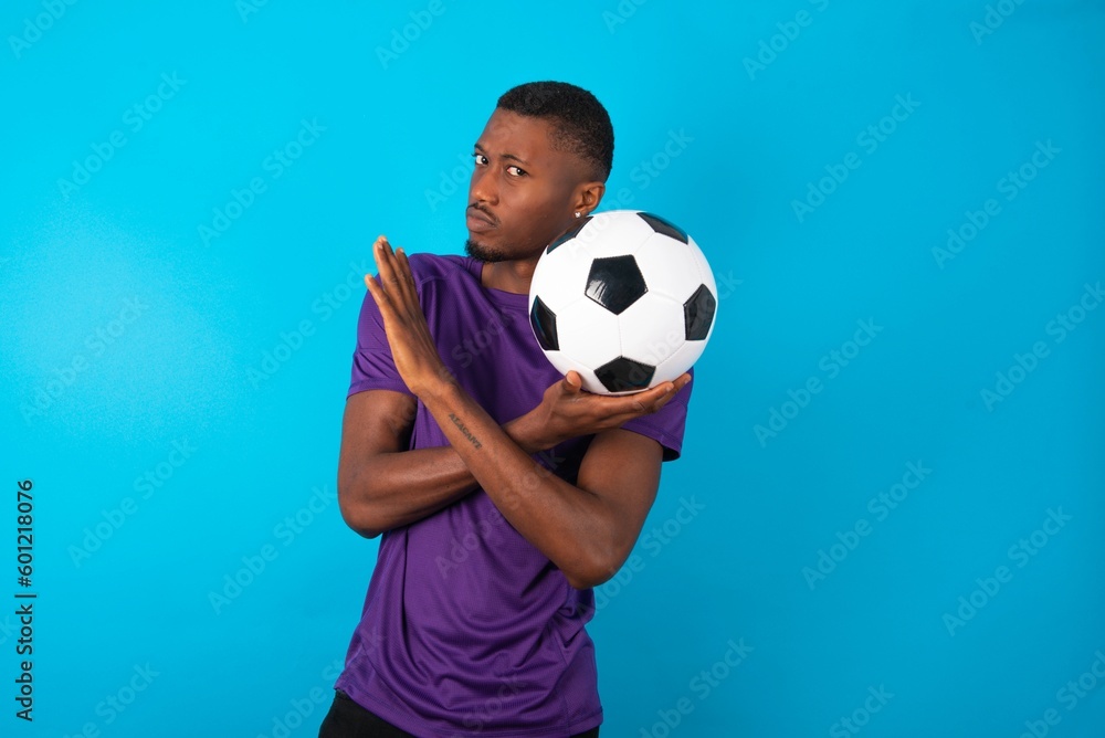 Man wearing purple T-shirt holding a ball over blue background Has rejection angry expression on face and crossing hands doing refusal negative sign. Man wearing purple T-shirt holding a ball over blue background Has rejection angry expression on face and crossing hands doing refusal negative sign.
