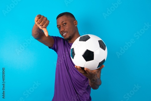 Discontent Man wearing purple T-shirt holding a ball over blue background shows disapproval sign, keeps thumb down, expresses dislike, frowns face in discontent. Negative feelings. © Jihan