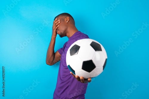 Man wearing purple T-shirt holding a ball over blue background covers eyes with palm and doing stop gesture, tries to hide. Don't look at me, I don't want to see, feels ashamed or scared.