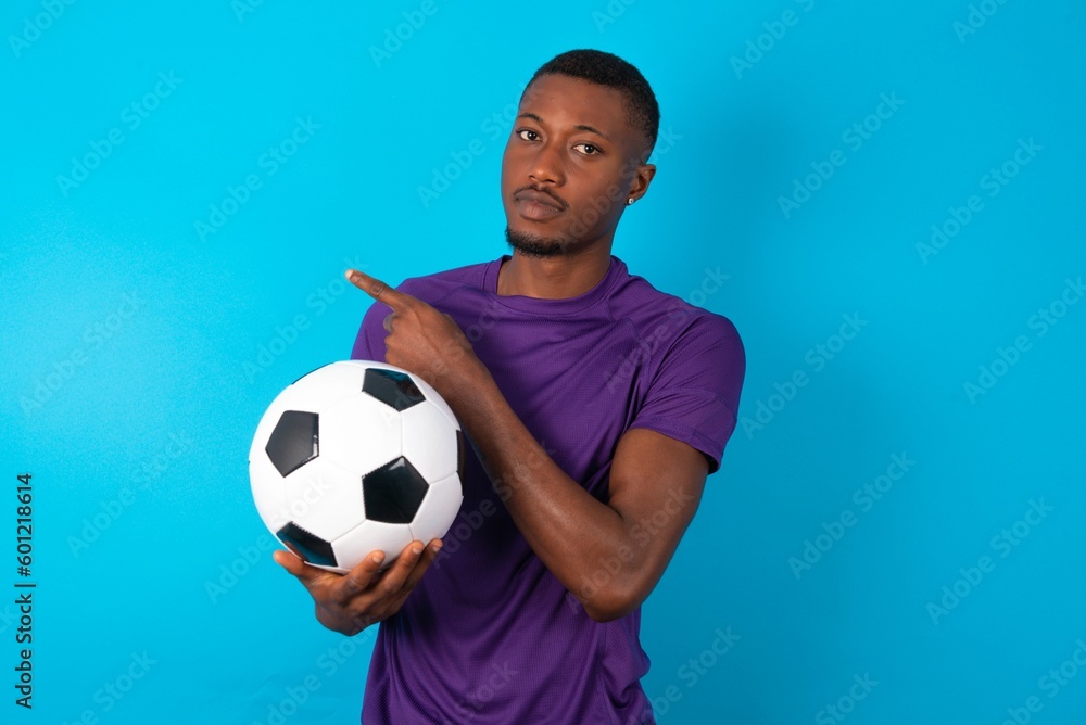 Man wearing purple T-shirt holding a ball over blue background smiling broadly at camera, pointing fingers away, showing something interesting and exciting. Man wearing purple T-shirt holding a ball over blue background smiling broadly at camera, pointing fingers away, showing something interesting and exciting.