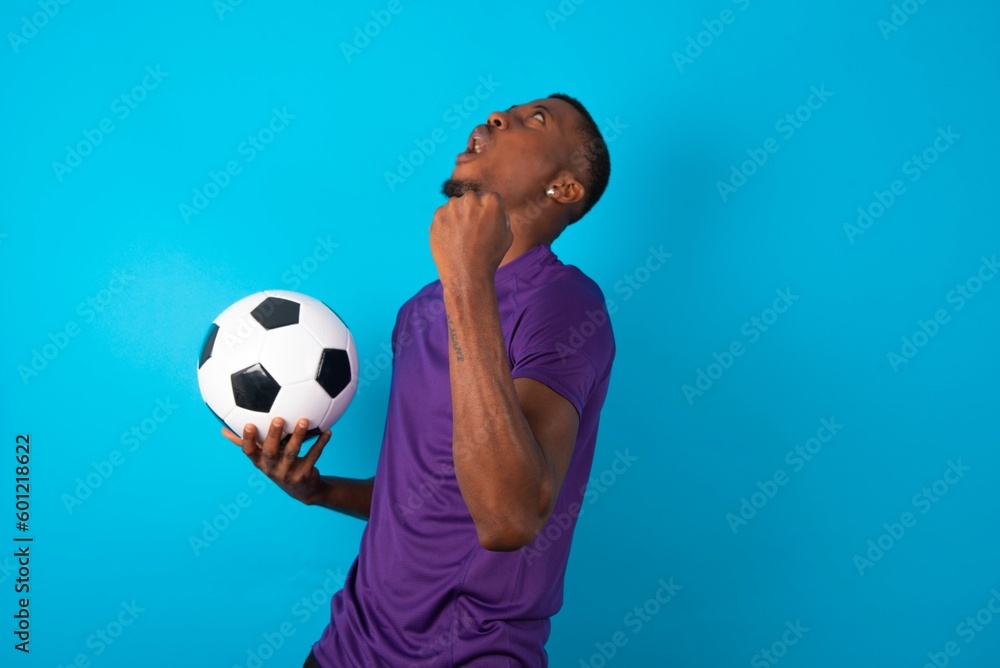 Attractive Man wearing purple T-shirt holding a ball over blue background celebrating a victory punching the air with his fists and a beaming toothy smile Attractive Man wearing purple T-shirt holding a ball over blue background celebrating a victory punching the air with his fists and a beaming toothy smile
