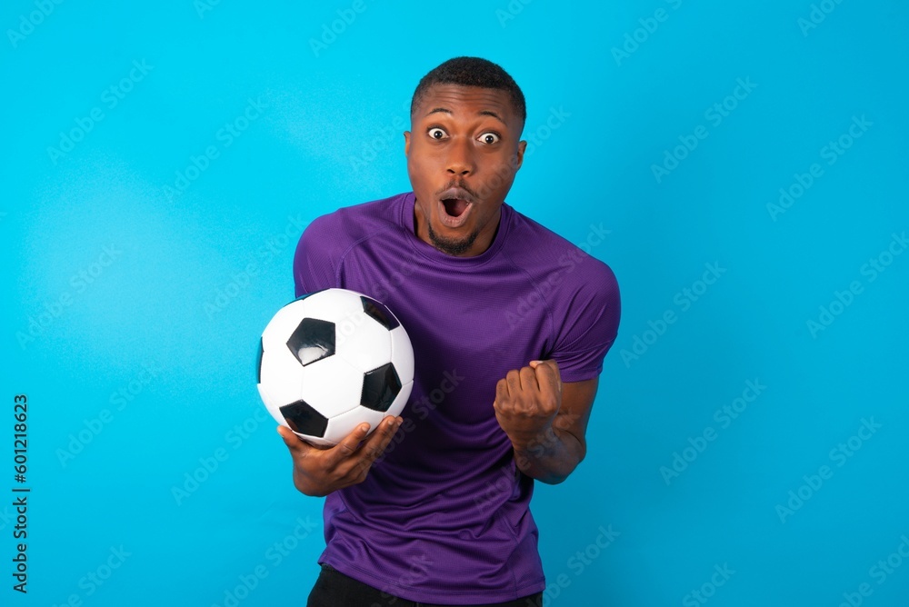 Joyful excited lucky Man wearing purple T-shirt holding a ball over blue  background cheering, celebrating success, screaming yes with clenched fists