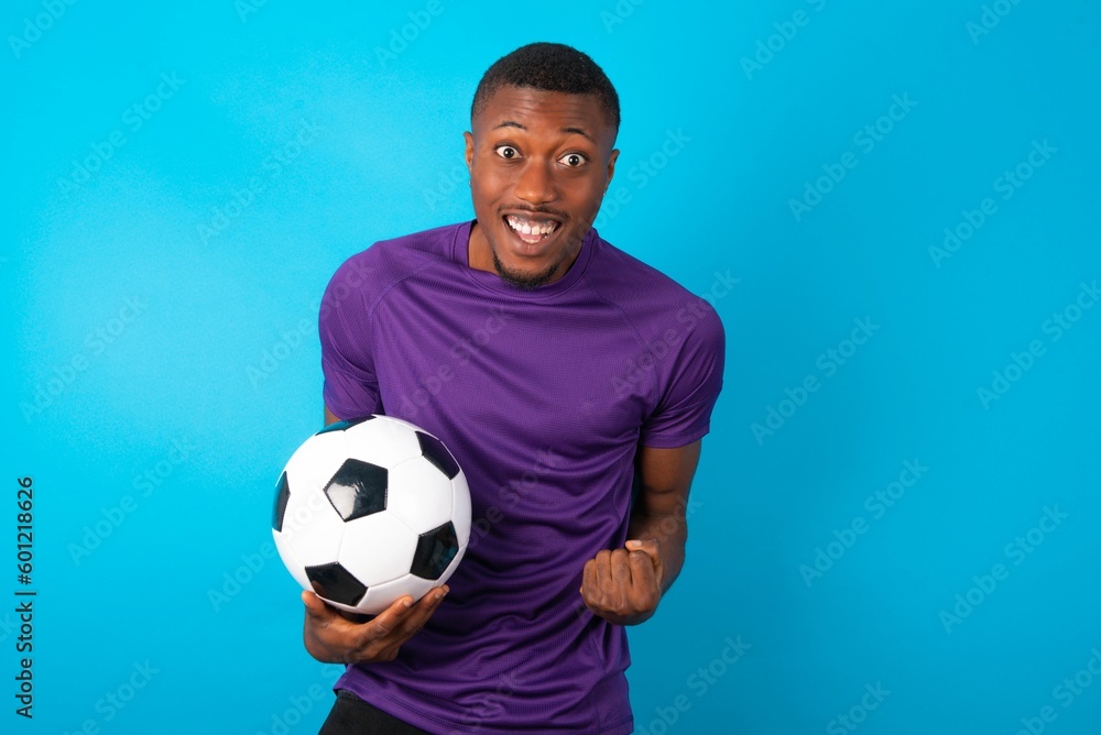 Man wearing purple T-shirt holding a ball over blue background raising fists up screaming with joy being happy to achieve goals. Man wearing purple T-shirt holding a ball over blue background raising fists up screaming with joy being happy to achieve goals.