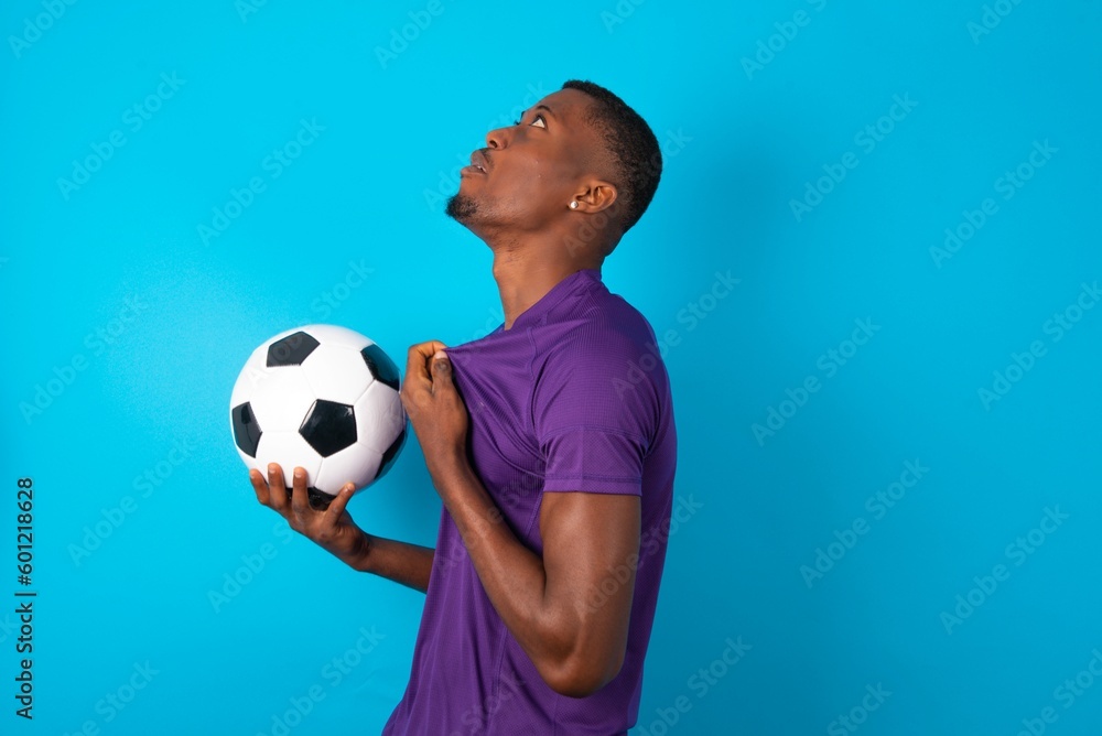 Man wearing purple T-shirt holding a ball over blue background excited and glad to achieve victory, clenches fists, screams in excitement with closed eyes,successful person.
