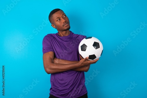 Image of upset Man wearing purple T-shirt holding a ball over blue background with arms crossed. Looking with disappointed expression aside after listening to bad news.