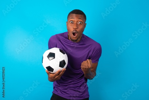 Joyful excited lucky Man wearing purple T-shirt holding a ball over blue background cheering, celebrating success, screaming yes with clenched fists