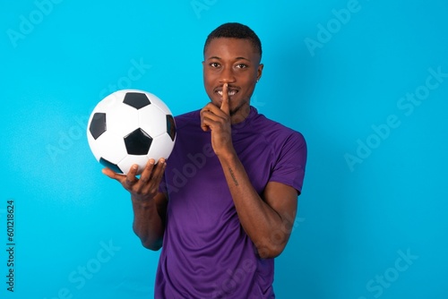 Smiling Man wearing purple T-shirt holding a ball over blue background makes shush gesture, holds fore finger over lips hides secret. Be mute, please.