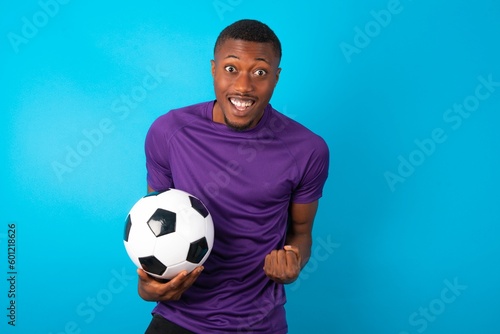 Man wearing purple T-shirt holding a ball over blue background raising fists up screaming with joy being happy to achieve goals.