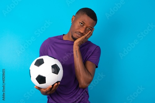 Very bored Man wearing purple T-shirt holding a ball over blue background holding hand on cheek while support it with another crossed hand, looking tired and sick.