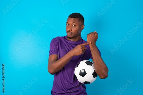 Man wearing purple T-shirt holding a ball over white background in hurry pointing to watch time, impatience, upset and angry for deadline delay