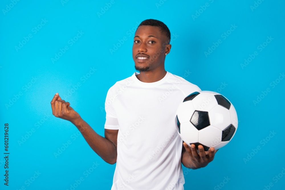 Young man wearing white T-shirt holding a ball over blue background , making money gesture. Young man wearing white T-shirt holding a ball over blue background , making money gesture.