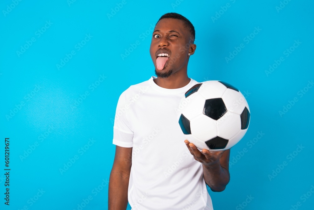 Young man wearing white T-shirt holding a ball over blue background with happy and funny face smiling and showing tongue.