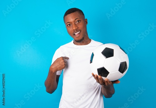 Young man wearing white T-shirt holding a ball over blue background being in stupor shocked, has astonished expression pointing at oneself with finger saying: Who me?