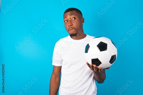 Amazed puzzled Young man wearing white T-shirt holding a ball over blue background , curves lips and has worried look, sees something awful in front.