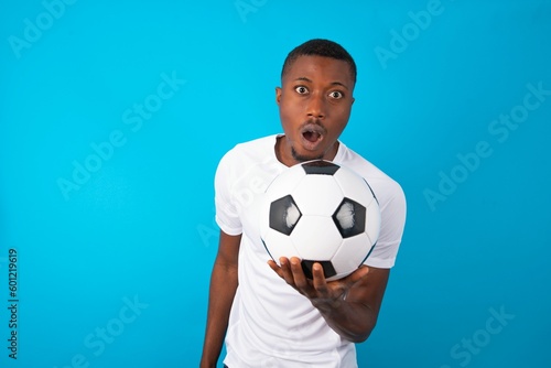 Young man wearing white T-shirt holding a ball over blue background having stunned and shocked look, with mouth open and jaw dropped exclaiming: Wow, I can't believe this. Surprise and shock