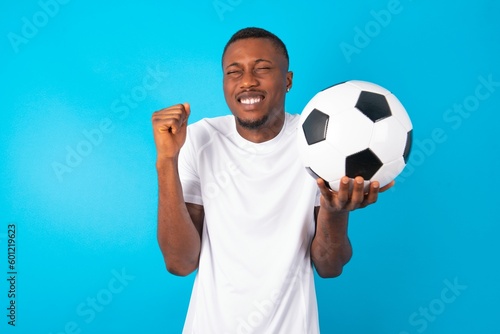 Young man wearing white T-shirt holding a ball over blue background rejoicing his success and victory clenching fists with joy being happy to achieve aim and goals. Positive emotions, feelings.