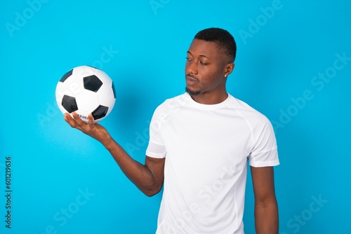 Young man wearing white T-shirt holding a ball over blue background with thoughtful expression, looks away keeps hands down bitting his lip thinks about something pleasant.