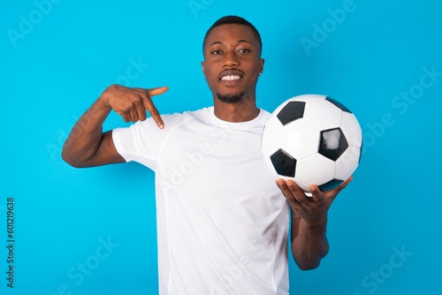 Young man wearing white T-shirt holding a ball over blue background with positive expression, points down with both index fingers, has broad interested smile. Look there, please.