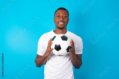Young man wearing white T-shirt holding a ball over blue background keeps palms together, has pleased expression. Glad attractive male makes request, pleads for mercy. Hopeful young adult.